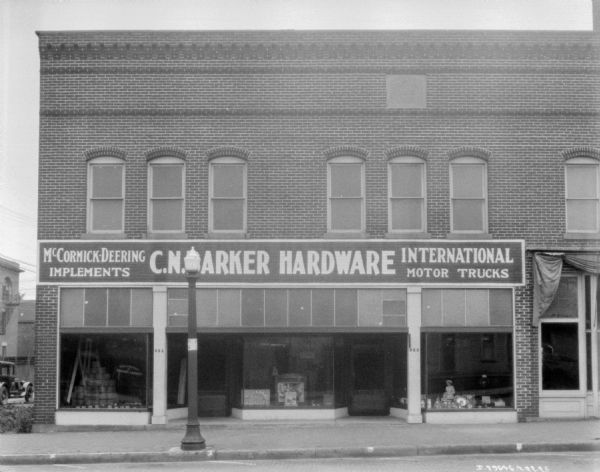View from street of the C.N. Barker hardware store and implement dealership. The brick building has large show windows, with twine and other items on display.