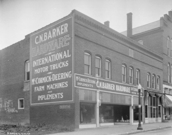 View from street towards the left corner and front of the C.N. Barker hardware store and implement dealership. Signs for the store are painted on the side of the building, and above the large show windows in the front along the sidewalk.