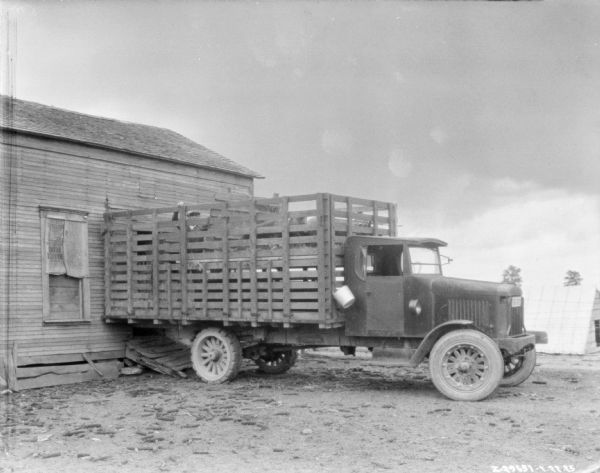 A man is standing in the back of a delivery truck. There are two cows, or cattle, in the back of the stake body bed of the truck. The truck is backed up to a farm building. There is another building in the background on the right.