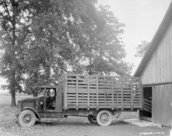 Left side view of a livestock delivery truck backed up to a doorway in a farm building. Cows or cattle are standing in the back of the truck which has a stake body. The driver's side door is open.