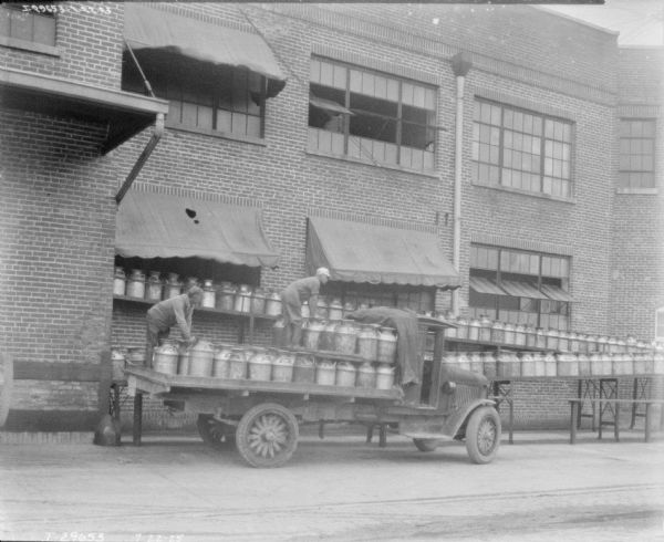 Men are loading milk cans into the back of a truck parked near a large brick building.