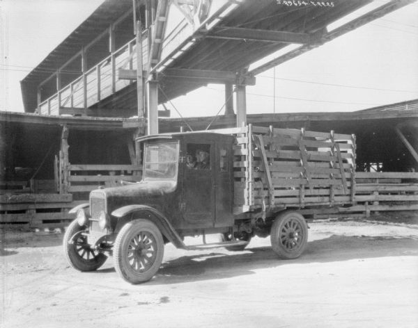 A man and woman are sitting in a livestock delivery truck. In the back of the truck, which has a stake body, is a cow. Behind them another man stands behind a fence in the background under an open shelter. Above the truck is a large, wide, covered wooden walkway. Perhaps a slaughterhouse with pens.