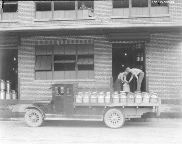 Two men are loading a truck with milk cans at a loading dock at a large brick building. A man is looking out of a second-story window above them