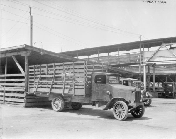 A truck with a stake body is transporting cattle at a cattle yard. The truck is backed up to the side of an open sided shed with fences on the side. Large wooden structures and other trucks are in the background.