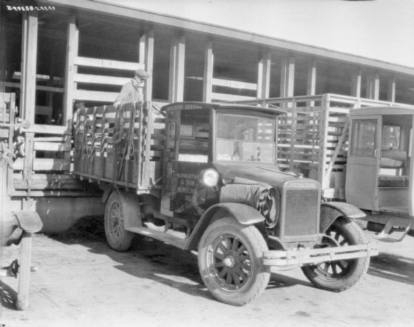 A man standing in the bed of a stake body truck transporting cattle. The truck is backed up to an open sided wood enclosure. On the side of the truck is a sign that reads: "Kenworthy & Son, Monrovia, Ind." Another truck is parked on the right.