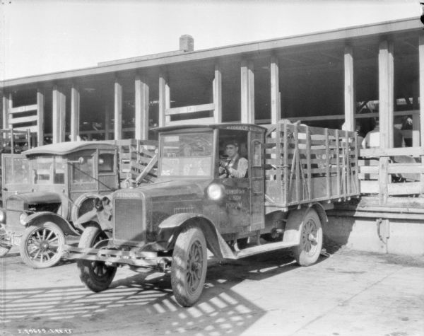 Three-quarter view from front left of a livestock delivery truck with stake body. There is a man in the driver's seat, and cattle in the bed of the truck. Behind the truck are other trucks, and they are all backed up against a wooden building with a roof and fence.