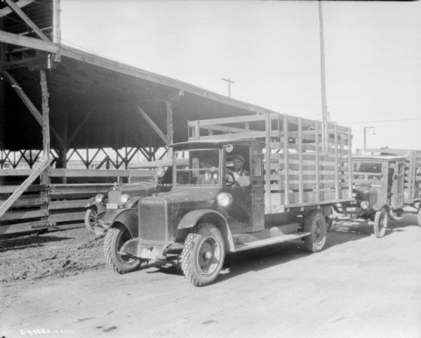 Two men are sitting in the cab of a livestock delivery truck with stake body. Large wooden structures and other trucks are in the background.