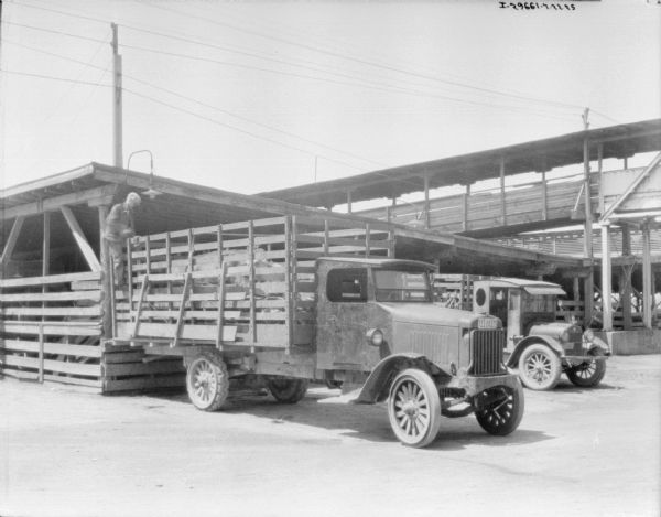 A man is standing at the back of a truck with a stake body. There are cattle standing in the bed of the truck, which is backed up to an open-sided building with a fence.