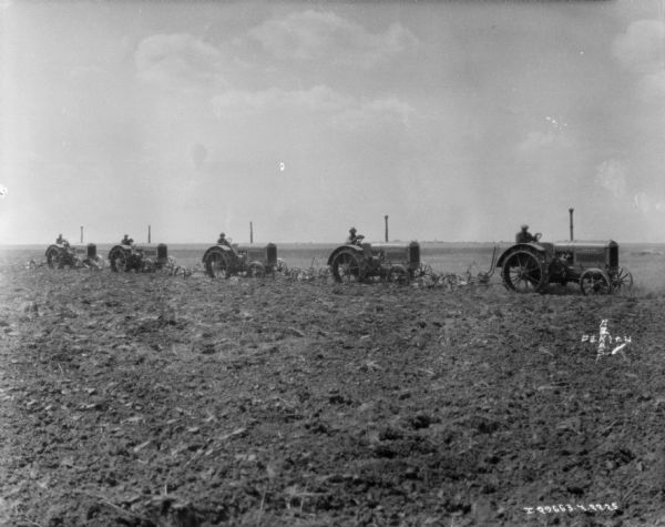 View across field towards five men in a line driving tractors to pull plows.