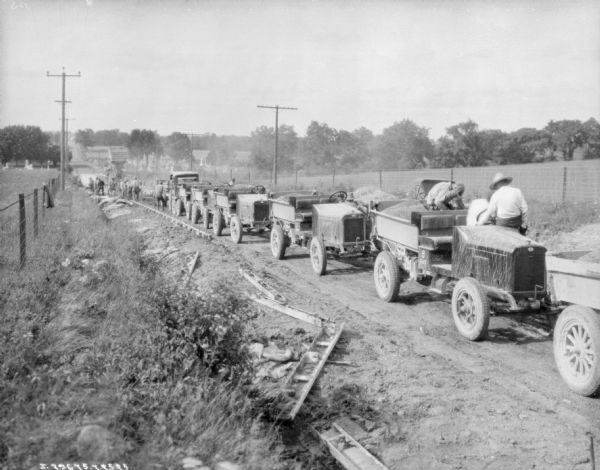 View from side of road of a line of trucks on a road. There is a large piece of machinery in the background where men are working. In the background is a cemetery.