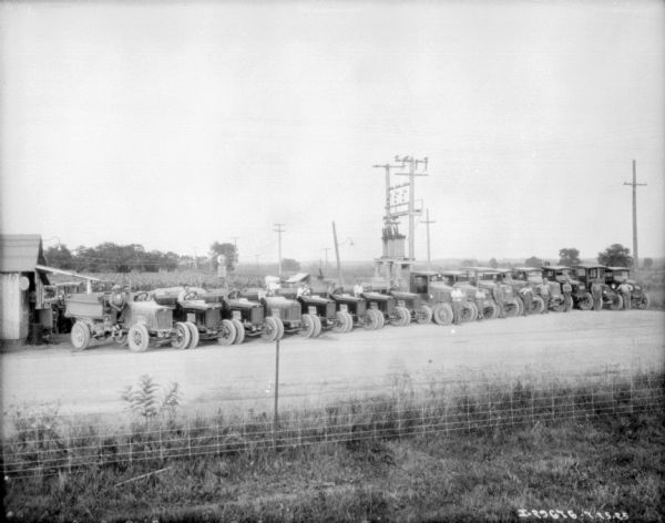 Slightly elevated view over fence towards a large group of men sitting or standing with trucks parked in a long line at an angle. In the background are fields.