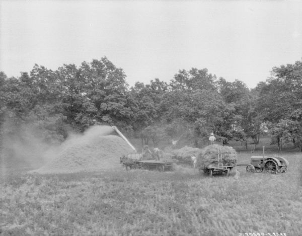 View across field towards a group of men with horse-drawn wagons using a thresher powered by a tractor. Trees are in the background.
