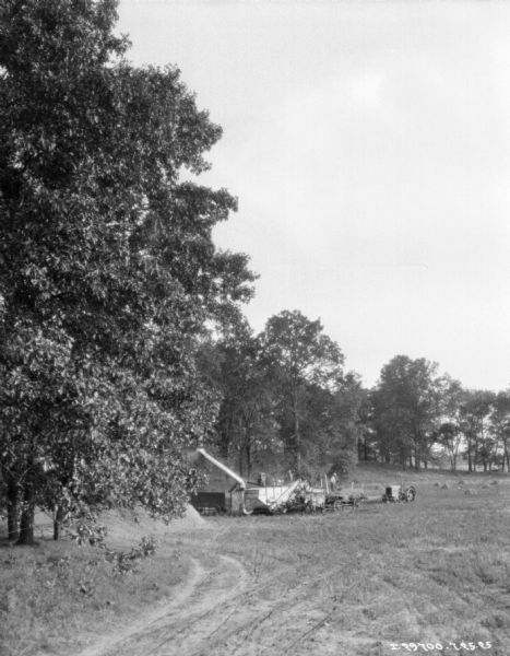 View across field towards men using a tractor-powered threshing machine. Some of the men are standing on horse-drawn wagson loaded with grain. The field is surrounded by trees.