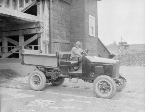 A man is sitting in a dump truck parked outdoors near an industrial building, perhaps at a quarry. The truck has a bench seat, and no cab enclosure.