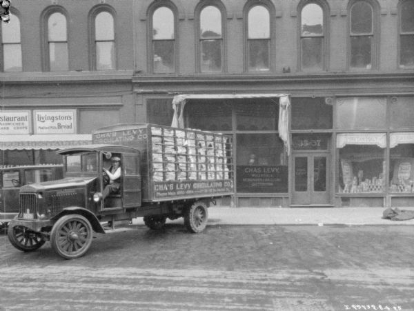 View across street towards a man sitting in the driver's seat of a delivery truck parked in front of the Chas. Levy storefront. The painted sign on the truck reads: "Cha's Levy Circulating Co."