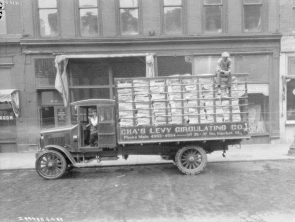 View across street towards a man sitting in the driver's seat of a fully loaded delivery truck parked at the curb in front of the Chas. Levy storefront. The painted sign on the truck reads: "Cha's Levy Circulating Co."