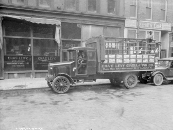 View across street towards two men sitting in the cab of a delivery truck parked at the curb in front of the Chas. Levy storefront. Another man is standing in the bed of the truck. The painted sign on the truck reads: "Cha's Levy Circulating Co."