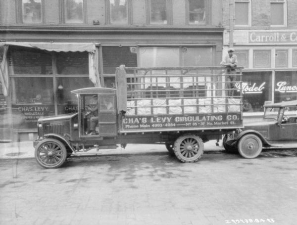 View across street towards two men sitting in the cab of a delivery truck parked at the curb in front of the Chas. Levy storefront. Another man is standing in the bed of the truck. The painted sign on the truck reads: "Cha's Levy Circulating Co."