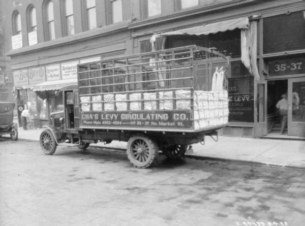 View across street towards a delivery truck parked at the curb in front of the Chas. Levy storefront. The painted sign on the truck reads: "Cha's Levy Circulating Co."