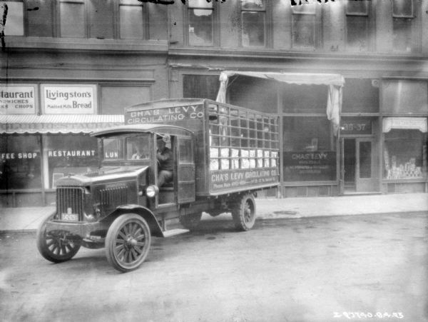 View across street towards two men sitting in the cab of a fully loaded delivery truck backed up at the curb in front of the Chas. Levy storefront. The painted sign on the truck reads: "Cha's Levy Circulating Co."