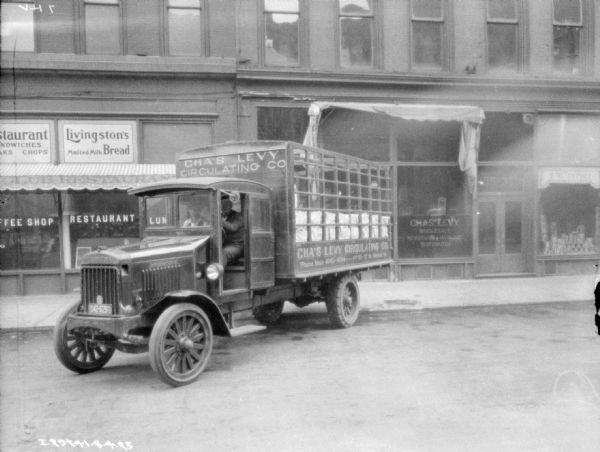 View across street towards two men sitting in the cab of a fully loaded delivery truck backed up at the curb in front of the Chas. Levy storefront. The painted sign on the truck reads: "Cha's Levy Circulating Co."