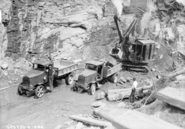 View looking down at men standing with trucks near a steam shovel working near a rock face.
