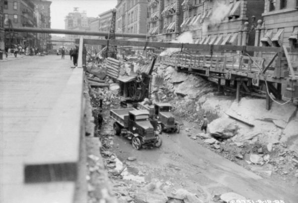 View along street towards pedestrians on the left. On the right, below street level, are construction workers standing below street level near trucks and a steam shovel. There are brick buildings lining both sides of the street.