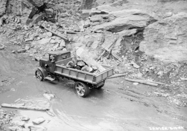 View looking down at a man driving a dump truck filled with large boulders through a construction site with puddles of water.