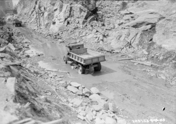 View looking down towards a man driving a dump truck through puddles of water at a construction site. In the background men are working among rocks and rock faces below street level.