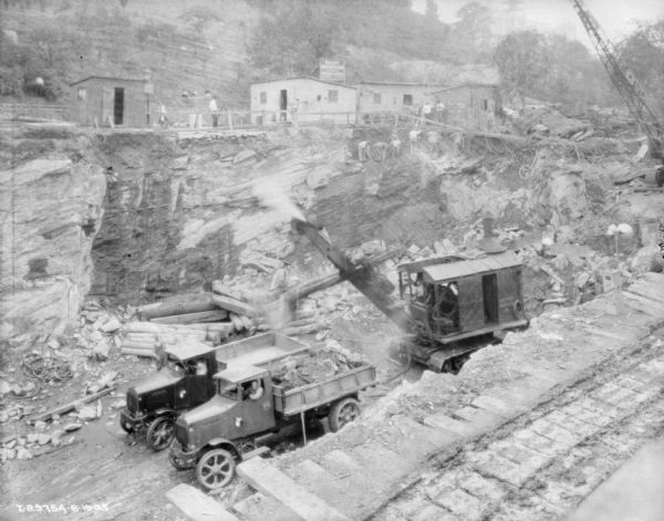Elevated view looking down at men driving dump trucks and operating a steam shovel and a crane below ground level at a construction site. On the opposite side of the site are fences and industrial buildings. There is a group of men working the exposed rock face just below the buildings. There is a hill with trees rising up in the background.