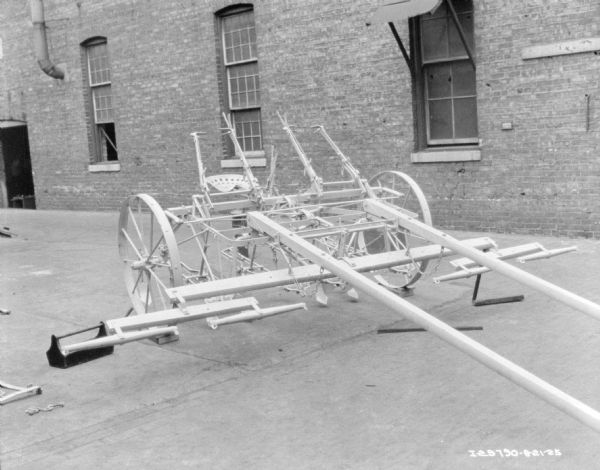 A two row cultivator displayed on the pavement in front of a brick factory building. A wood toolbox is on the ground on the left.