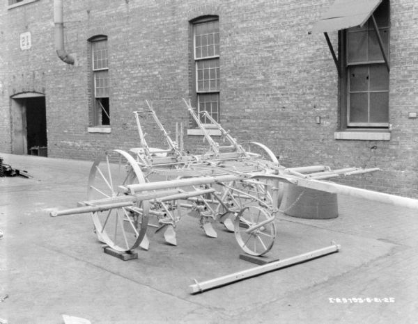 A two row cultivator displayed on the pavement in front of a brick factory building.