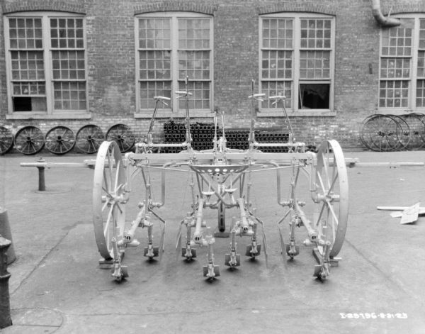 A two row cultivator displayed on the pavement in front of a brick factory building. Parts and wheels are stacked along the brick factory wall. 