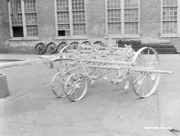 A two row cultivator displayed on the pavement in front of a brick factory building. Parts and wheels are stacked along the brick factory wall.