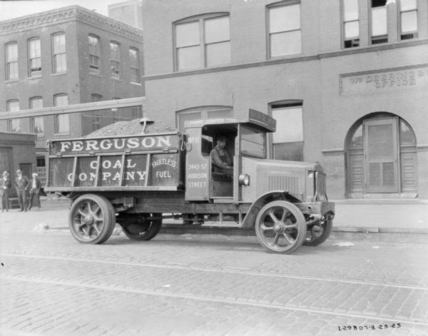 View across street toward a man sitting in a Ferguson Coal Company delivery truck. A sign on the brick building in the background reads: "Wm Deering & __ Office." Three men are standing in the background on the left.