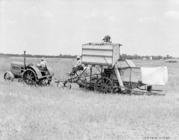 A man is driving a tractor to pull a man sitting on a harvester thresher in a field.