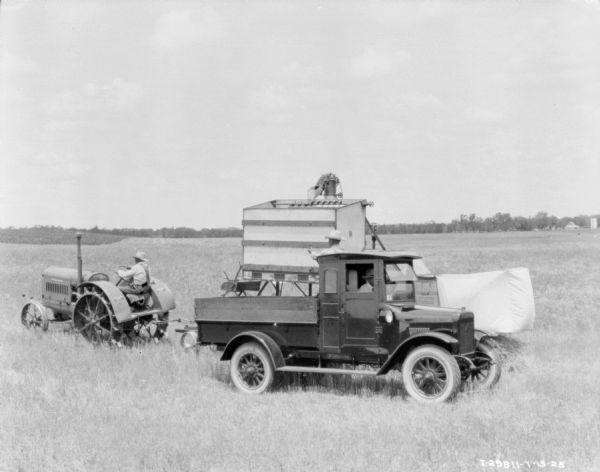 A man in a truck is parked in front of a man driving a tractor to pull a harvester thresher. Another man is standing behind the truck.
