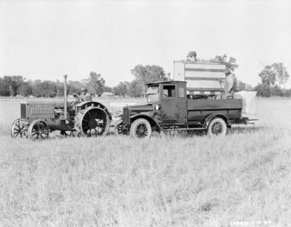 A man is standing in the back of an International Harvester truck parked in a field in front of a harvester thresher. A man on a McCormick-Deering tractor is pulling the harvester thresher.