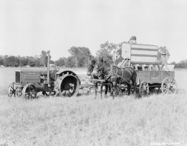 Two boys are sitting on a horse-drawn wagon in front of a tractor-drawn harvester thresher in a field.