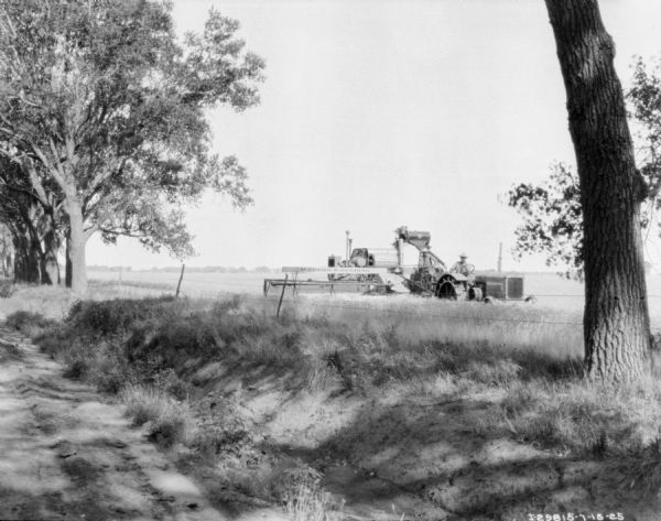 View from road towards a man driving a tractor to pull a harvester thresher in a field.