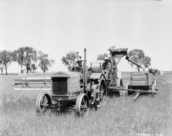 Three-quarter view from front left of a man driving a McCormick-Deering tractor to pull a man on a harvester thresher in a field.