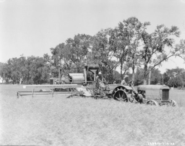 Three-quarter view from front right of a man driving a tractor to pull a man on a harvester thresher in a field.