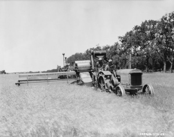 Three-quarter view from front right of a man driving a McCormick-Deering tractor to pull a man on a harvester thresher in a field.