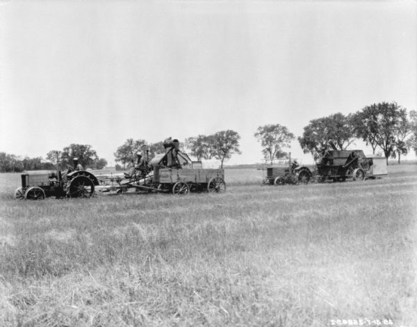 View across field towards two tractor-drawn harvester threshers. Each one is drawn by a man driving a tractor, and a man is sitting on each harvester thresher. There is a wagon next to the thresher on the left.