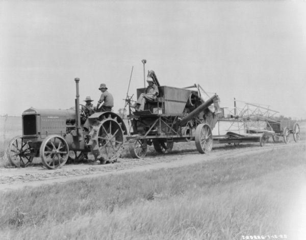 Three men are sitting on a tractor and a harvester thresher parked in a field. In the background is a wagon.