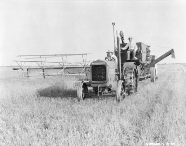 Three-quarter view from front left of a man driving a McCormick-Deering tractor to pull a man sitting on a harvester thresher in a field.