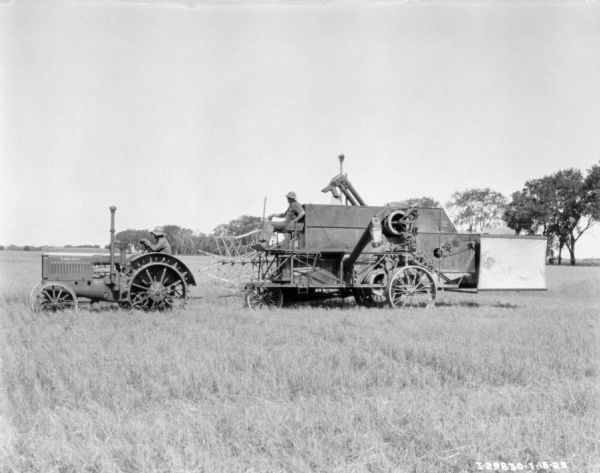 Profile view of left side of a man driving a McCormick-Deering tractor to pull a man on a harvester thresher in a field.
