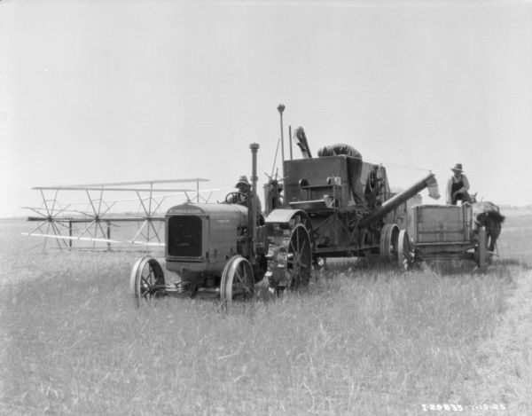 View from front of a man driving a tractor pulling a McCormick-Deering harvester thresher. A man is standing on the thresher looking down at something inside, and another man is standing on a horse-drawn wagon on the right.