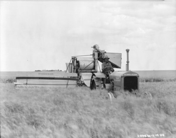 View from front of a man using a McCormick-Deering tractor to pull a harvester thresher in a field.