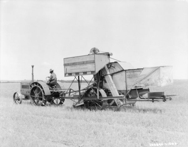 Three-quarter view from left rear of a man driving a tractor pulling a McCormick-Deering No. 8 power drive harvester thresher.